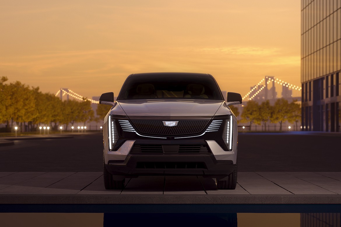 Front view of 2025 Cadillac Escalade IQ at dusk with LED lights reflected in water, set in modern cityscape.