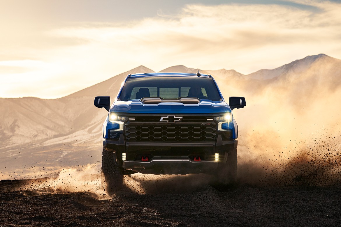 Front view of 2025 Chevrolet Silverado ZR2 kicking up dust off-road at sunset with mountains in background.